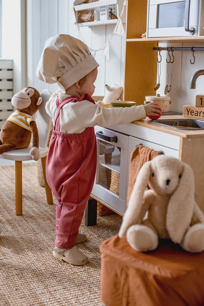 Home Adorable child wearing a chef hat playing in a toy kitchen with stuffed animals.
